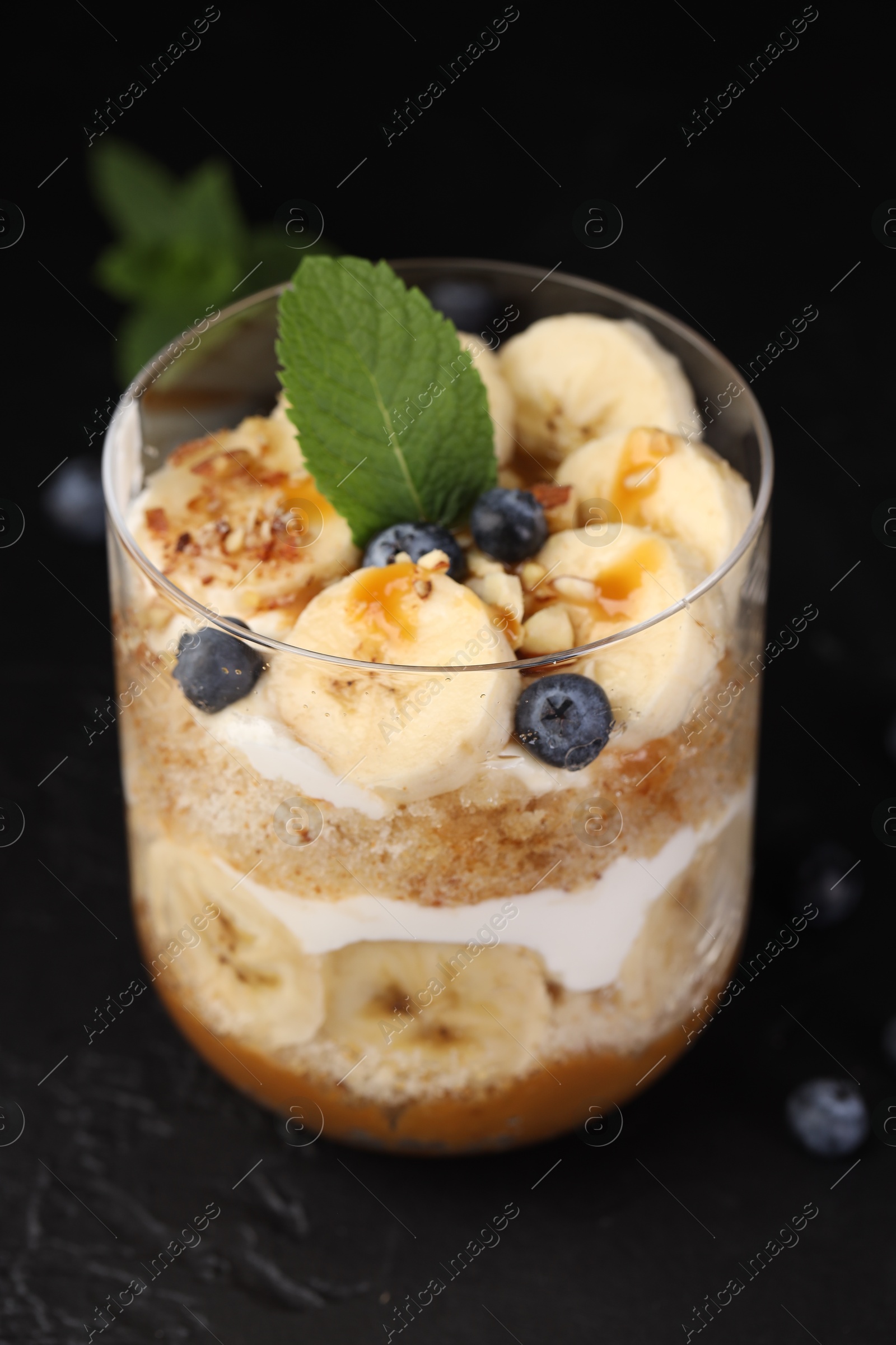 Photo of Tasty trifle dessert. Sponge cake, blueberries, banana and whipped cream in glass on black table, closeup