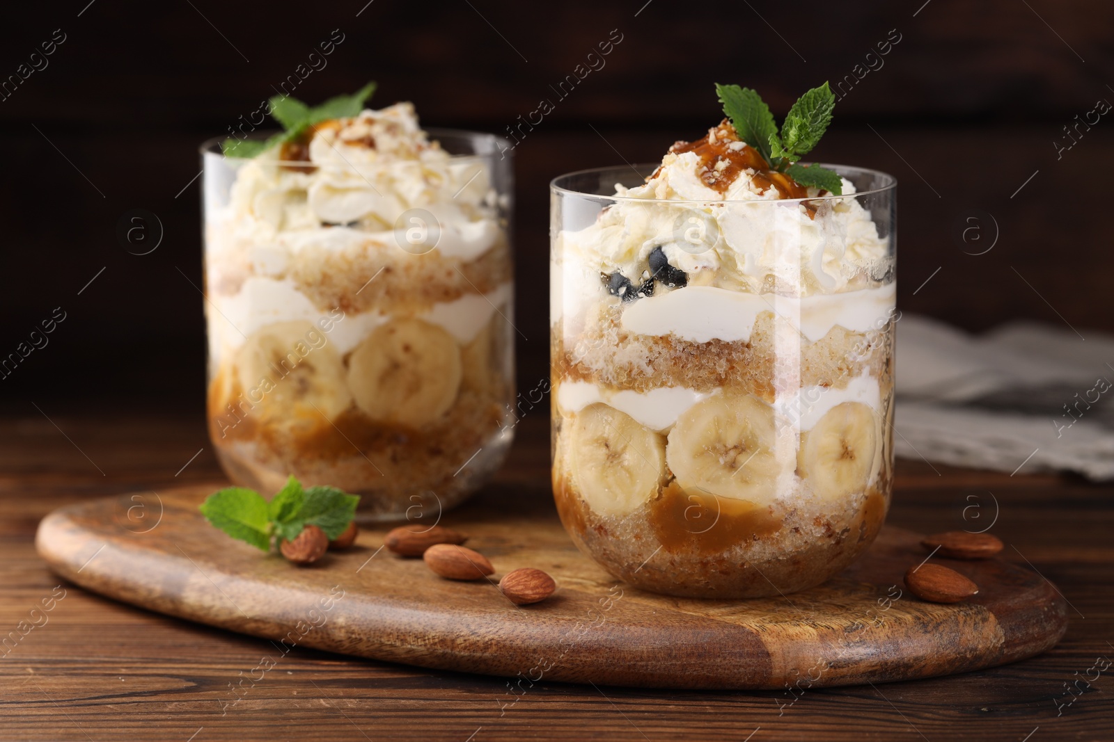 Photo of Tasty trifle dessert. Sponge cake, banana and whipped cream in glasses on wooden table, closeup