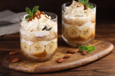 Photo of Tasty trifle dessert. Sponge cake, banana and whipped cream in glasses on wooden table, closeup