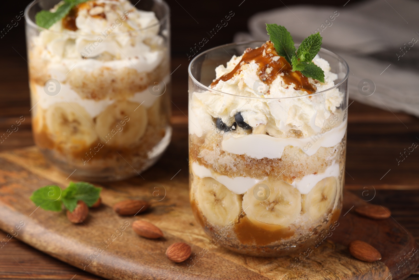 Photo of Tasty trifle dessert. Sponge cake, banana and whipped cream in glasses on wooden table, closeup