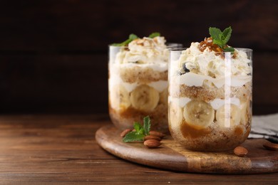 Photo of Tasty trifle dessert. Sponge cake, banana and whipped cream in glasses on wooden table, space for text