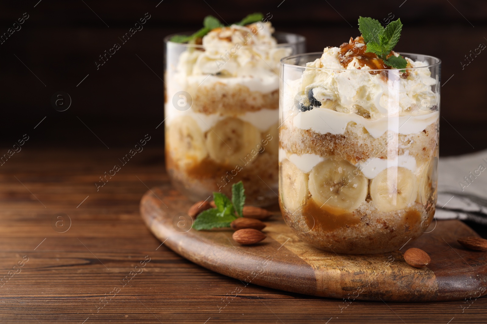 Photo of Tasty trifle dessert. Sponge cake, banana and whipped cream in glasses on wooden table, closeup