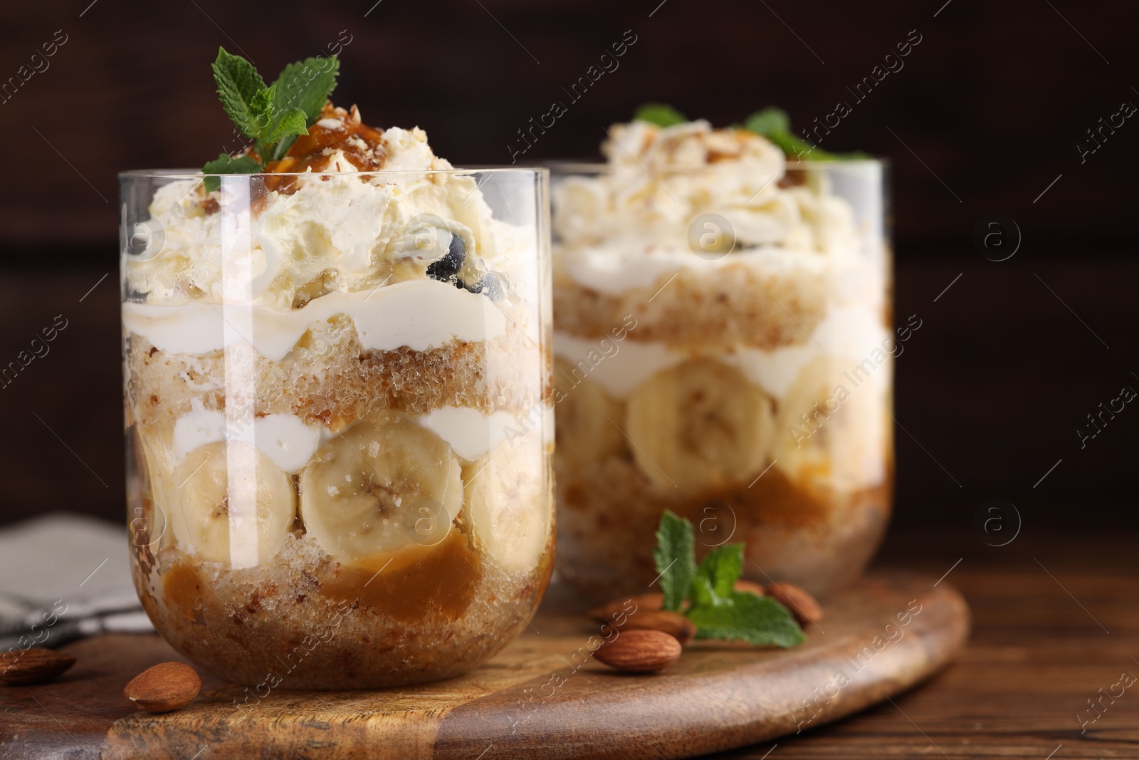 Photo of Tasty trifle dessert. Sponge cake, banana and whipped cream in glasses on wooden table, closeup