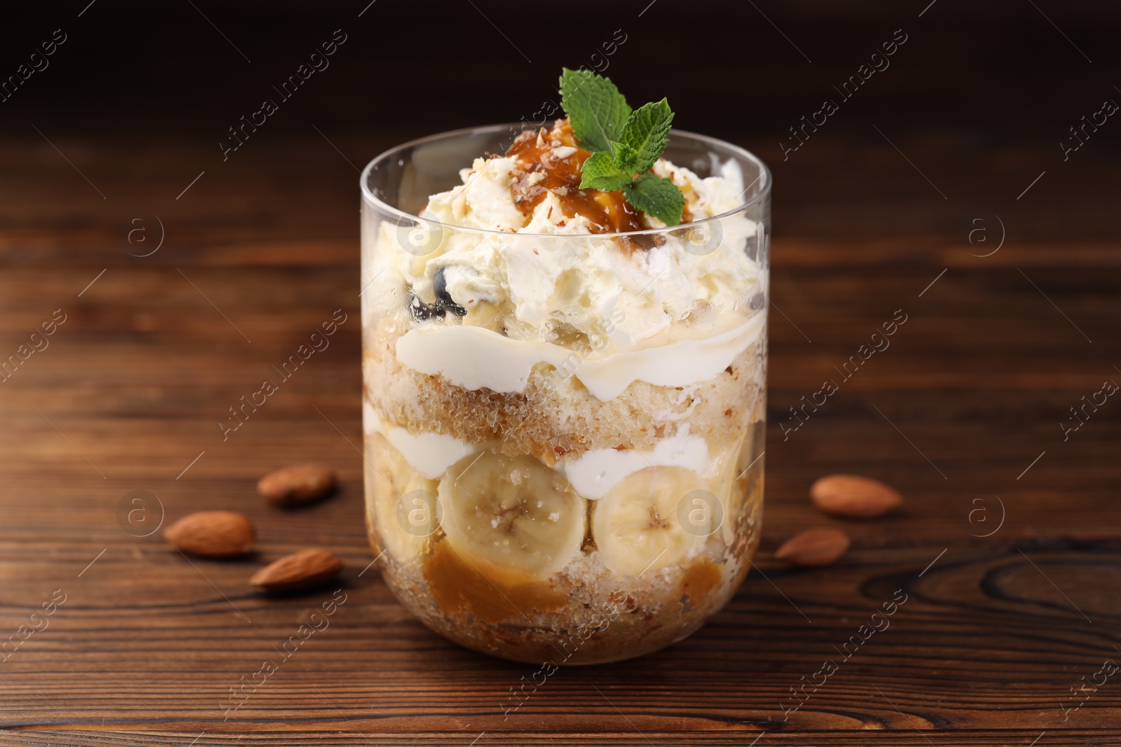 Photo of Tasty trifle dessert. Sponge cake, banana and whipped cream in glass on wooden table, closeup