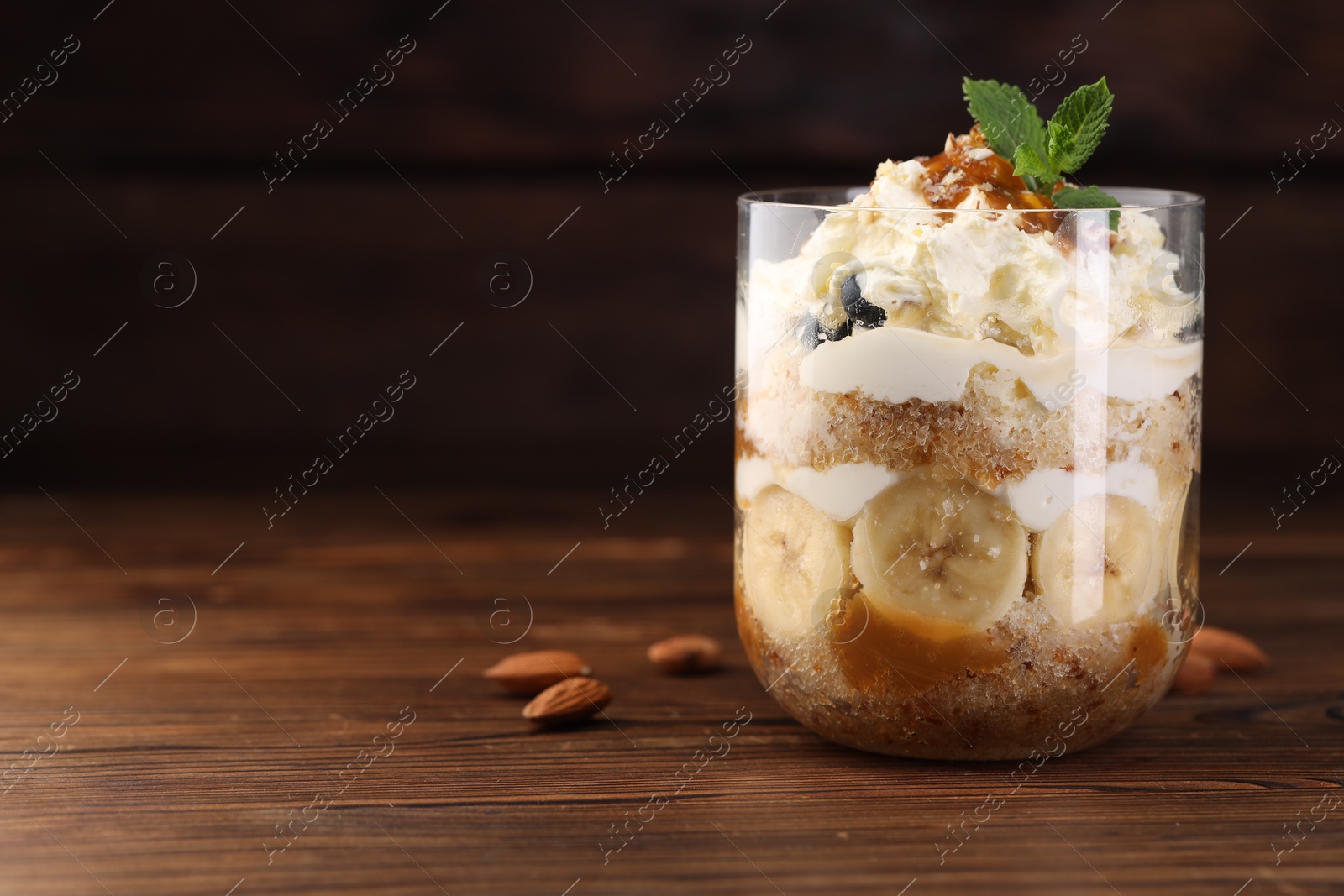 Photo of Tasty trifle dessert. Sponge cake, banana and whipped cream in glass on wooden table, closeup. Space for text