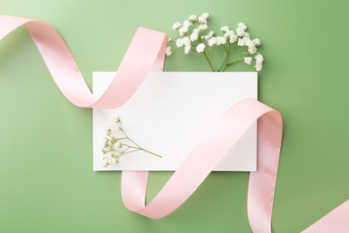 Photo of Blank card, ribbon and gypsophila flowers on green background, top view