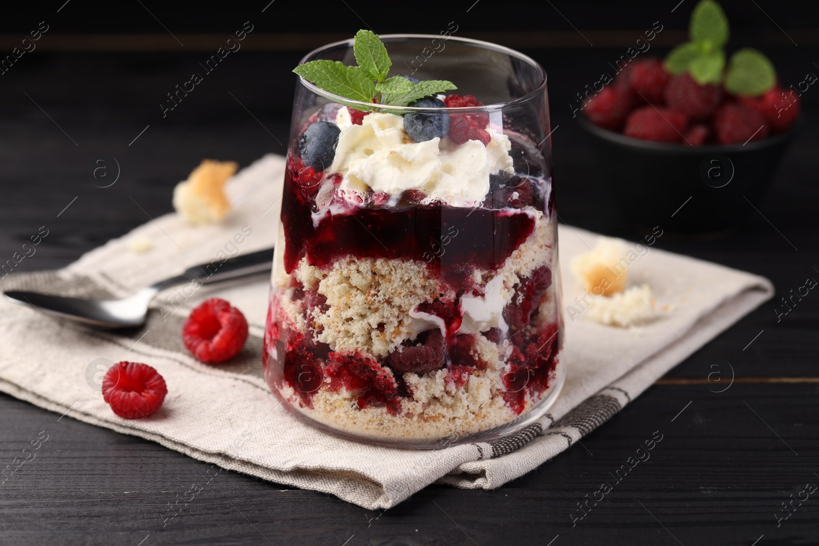 Photo of Tasty trifle dessert. Berries, sponge cake and whipped cream in glass on black wooden table