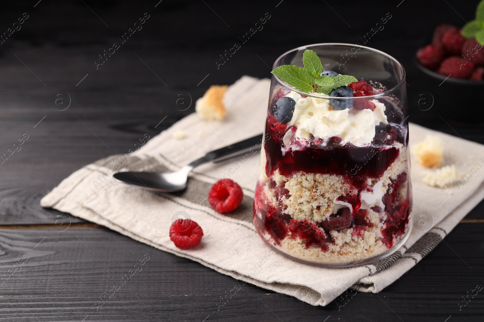 Photo of Tasty trifle dessert. Berries, sponge cake and whipped cream in glass on black wooden table, space for text