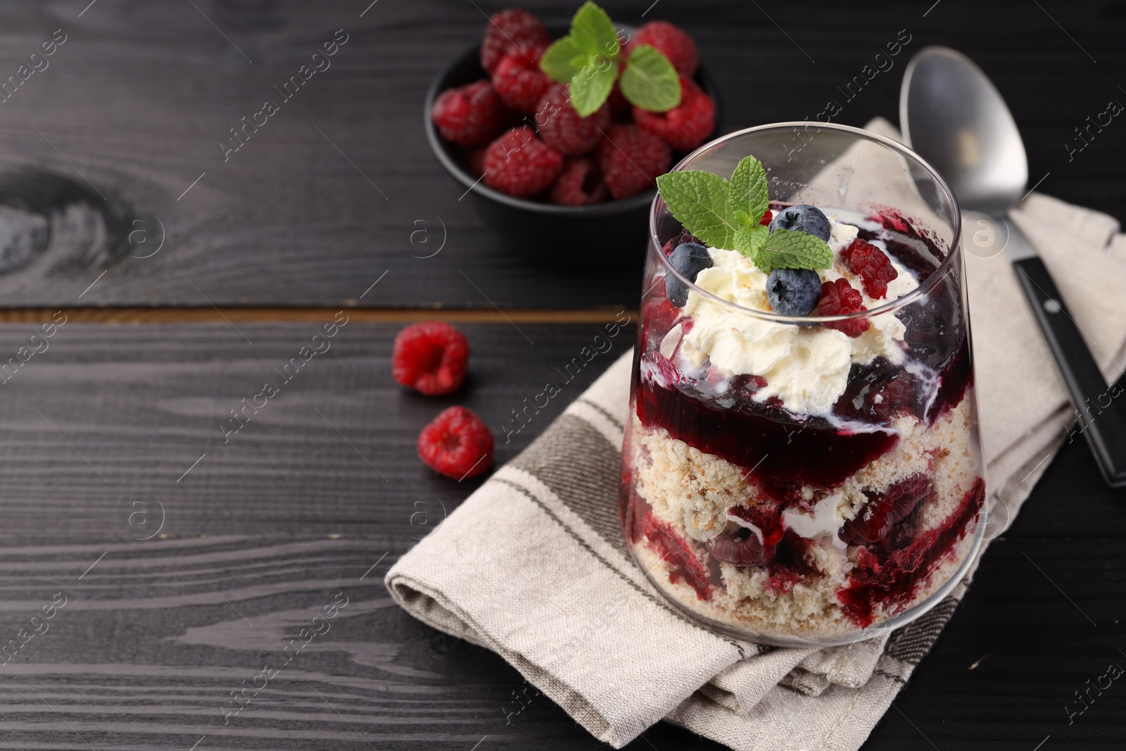 Photo of Tasty trifle dessert. Berries, sponge cake and whipped cream in glass on black wooden table, space for text