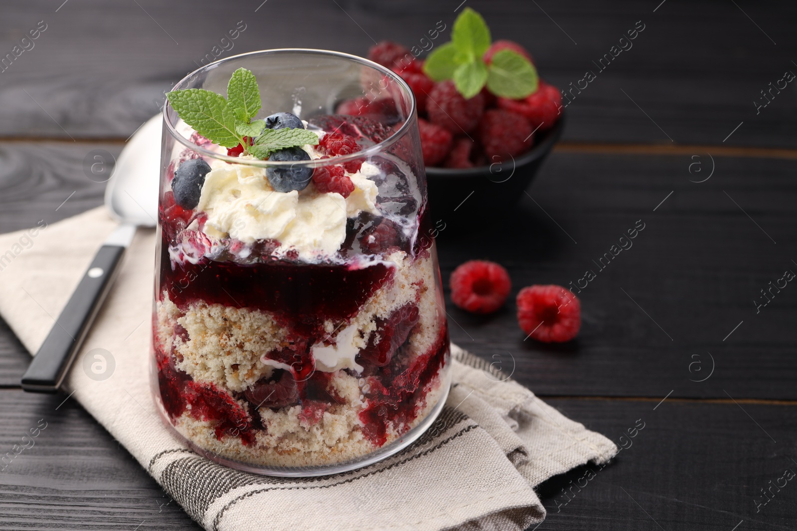 Photo of Tasty trifle dessert. Berries, sponge cake and whipped cream in glass on black wooden table, space for text