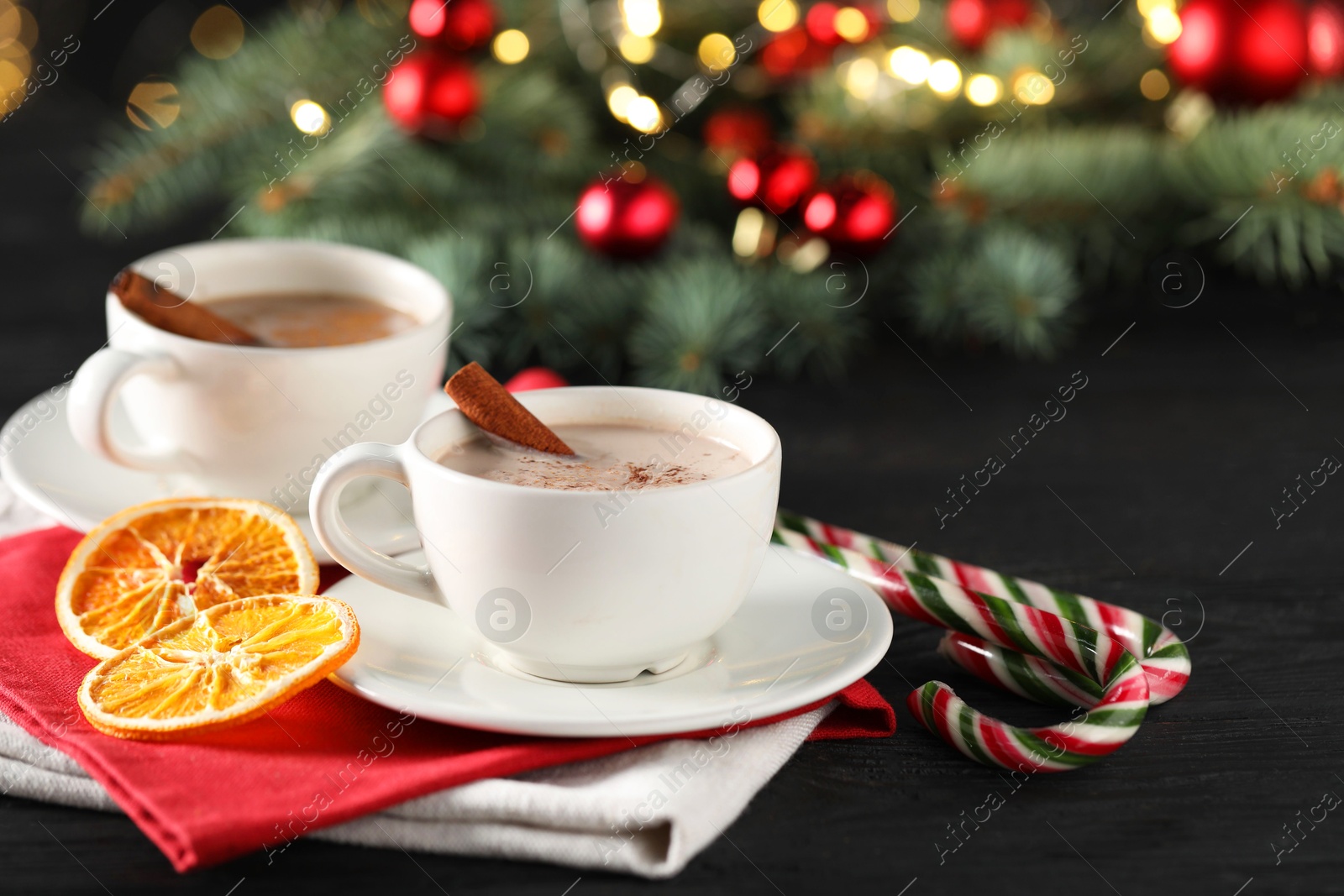 Photo of Tasty hot cocoa drink with cinnamon sticks in cups, dry orange and candy canes on black wooden table