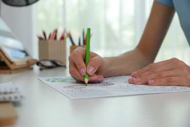 Young woman coloring antistress page at desk indoors, closeup