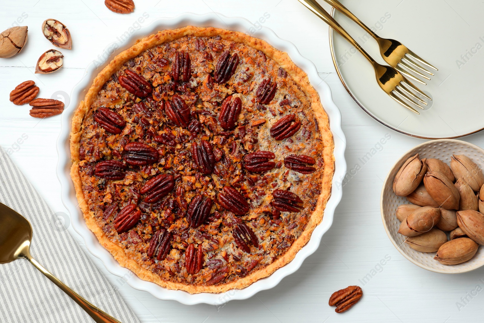 Photo of Delicious pecan pie in baking dish, fresh nuts and cake server on white wooden table, flat lay