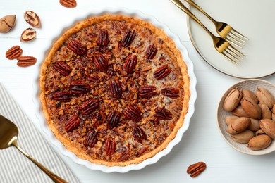 Photo of Delicious pecan pie in baking dish, fresh nuts and cake server on white wooden table, flat lay