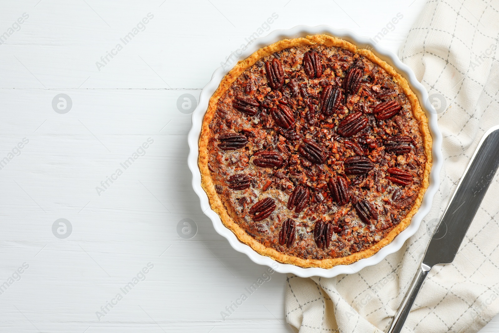 Photo of Delicious pecan pie in baking dish and knife on white wooden table, flat lay. Space for text