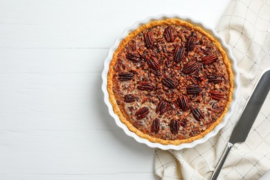 Photo of Delicious pecan pie in baking dish and knife on white wooden table, flat lay. Space for text