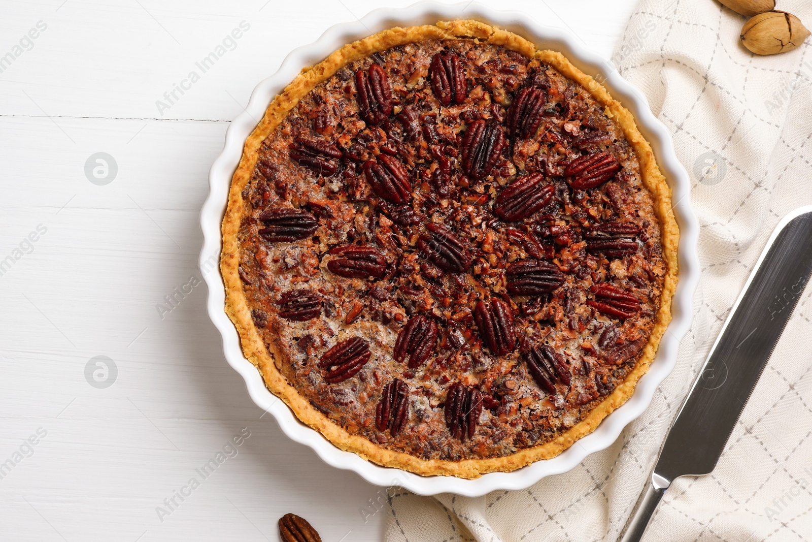 Photo of Delicious pecan pie in baking dish and knife on white wooden table, flat lay