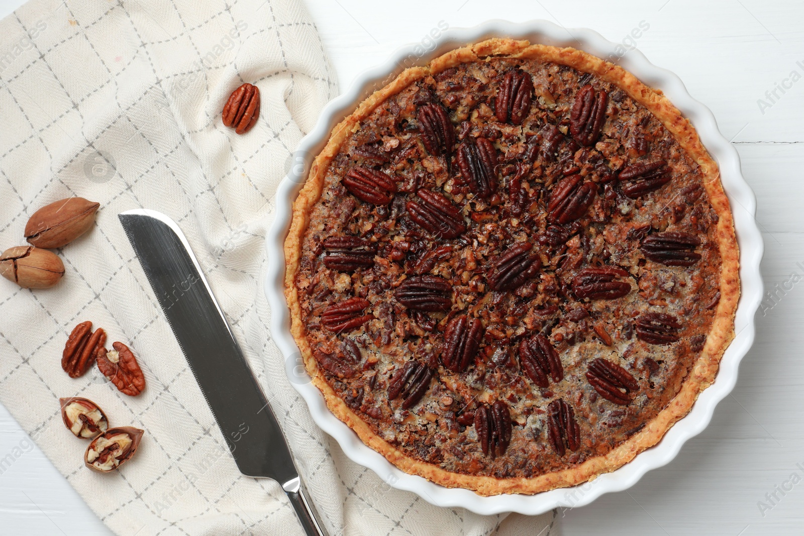 Photo of Delicious pecan pie in baking dish, knife and fresh nuts on white wooden table, flat lay