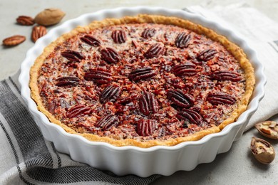 Photo of Delicious pecan pie in baking dish and fresh nuts on gray textured table, closeup