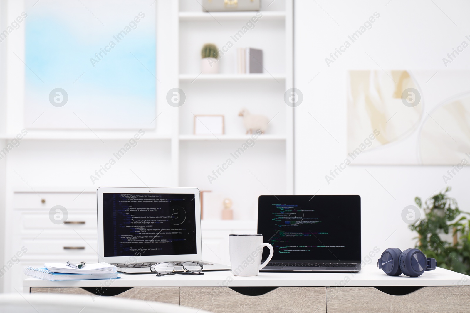 Photo of Programmer's workplace. Modern laptops with unfinished code, cup and other supplies on desk indoors