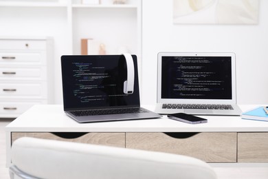 Photo of Programmer's workplace. Modern laptops with unfinished code, headphones and smartphone on desk indoors