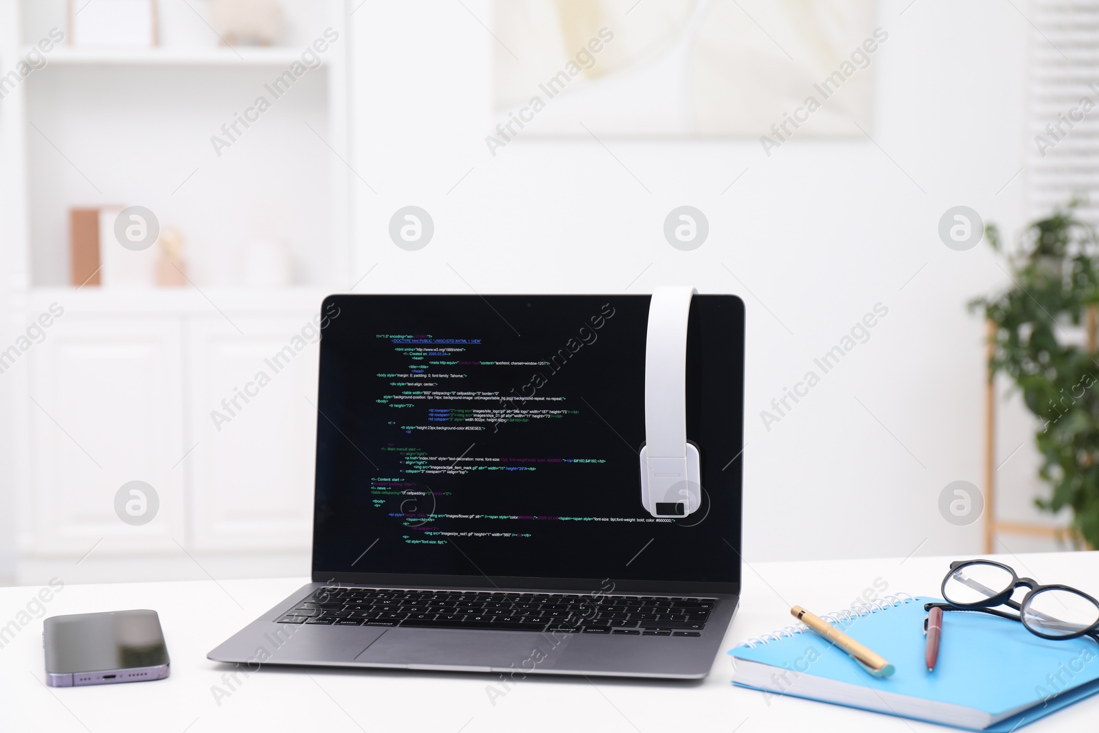 Photo of Programmer's workplace. Modern laptop with unfinished code, headphones and other supplies on white desk indoors