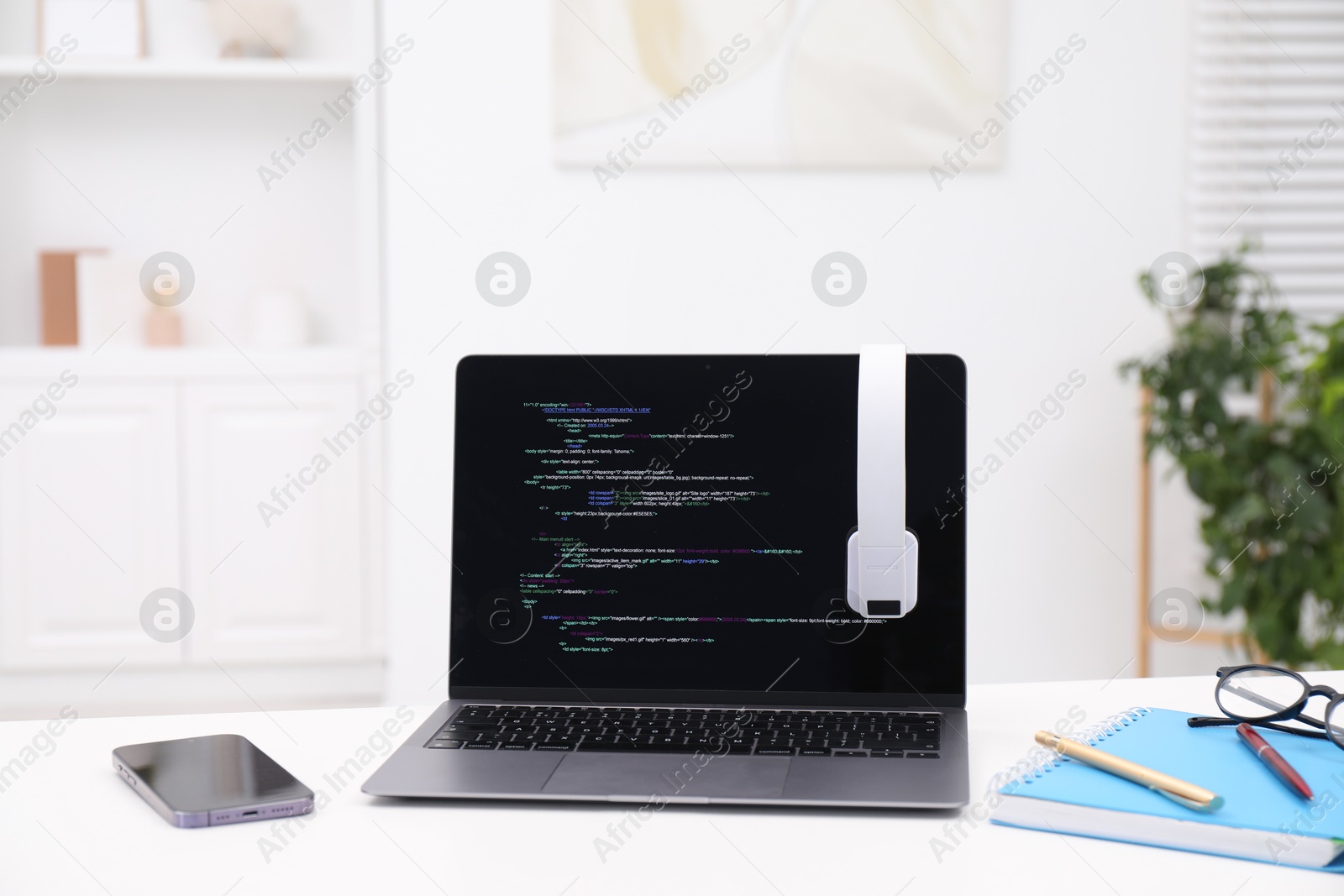 Photo of Programmer's workplace. Modern laptop with unfinished code, headphones and other supplies on white desk indoors