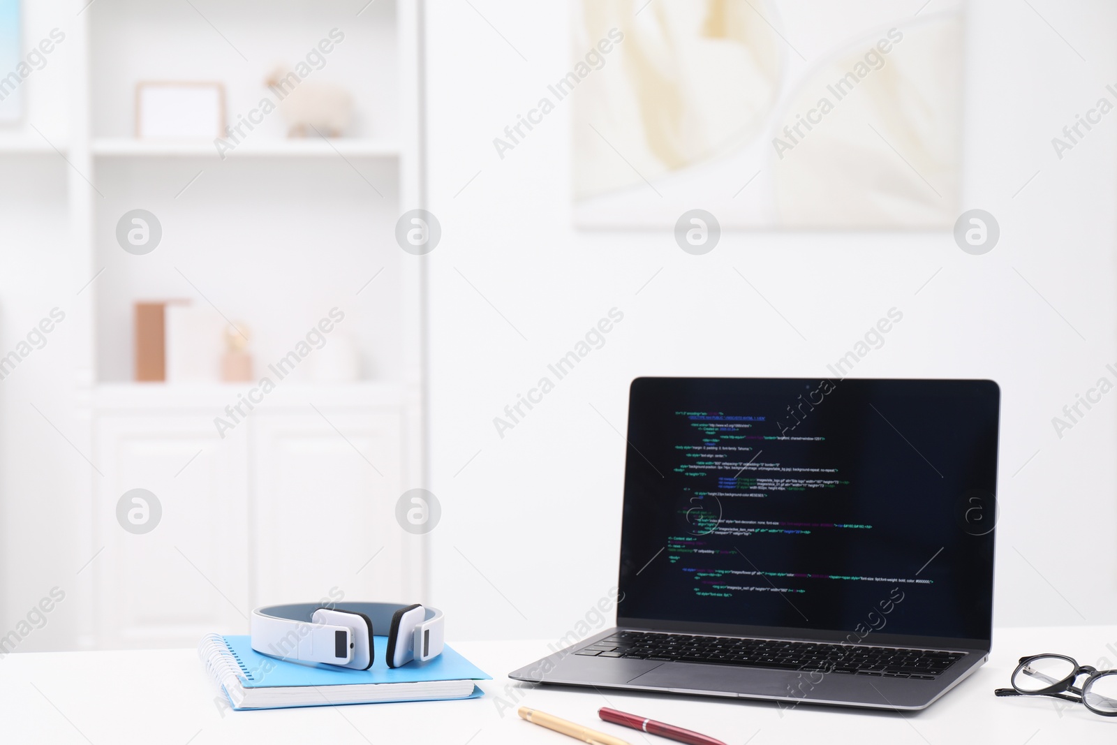 Photo of Programmer's workplace. Modern laptop with unfinished code, headphones and other supplies on white desk indoors