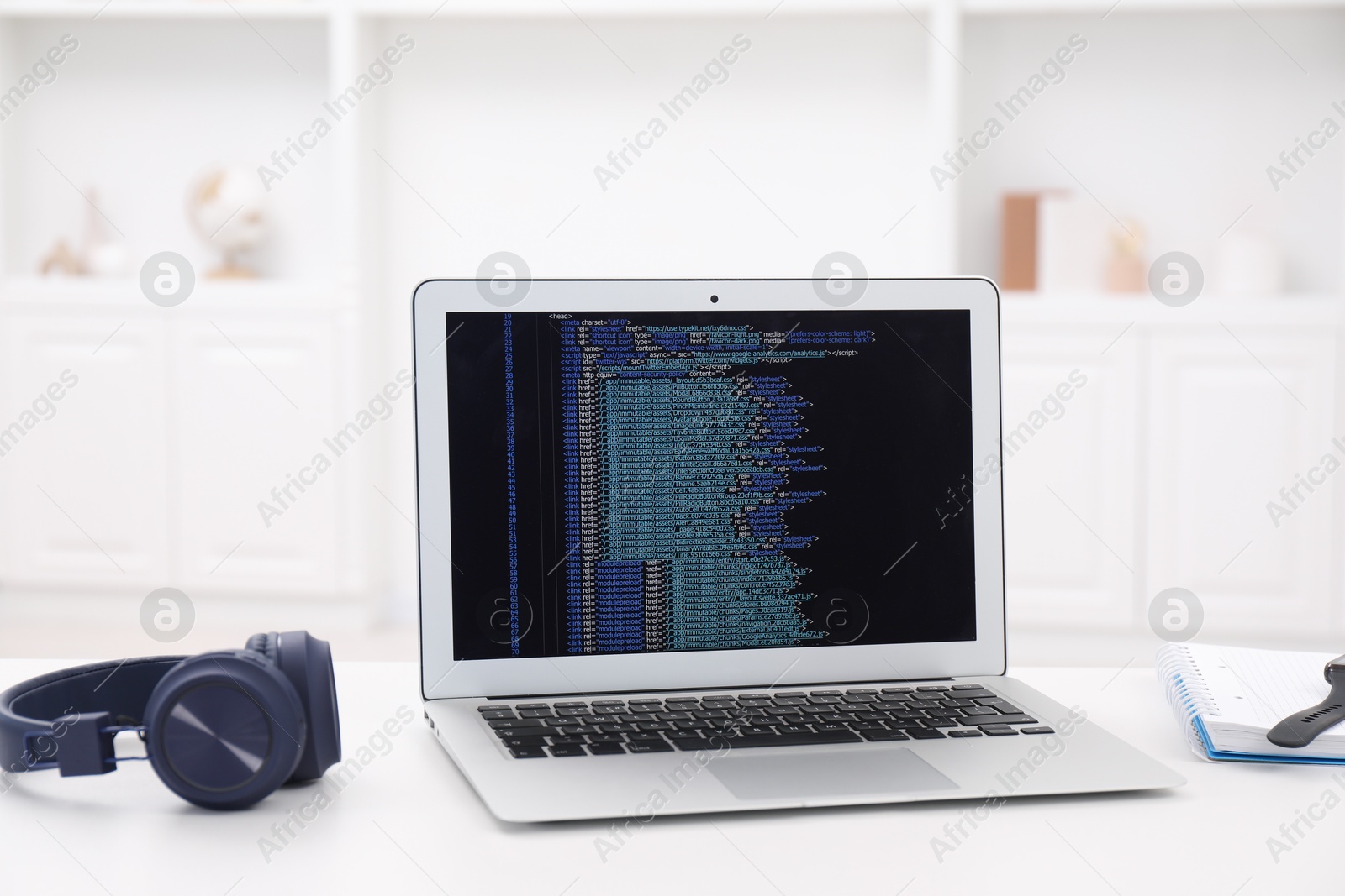 Photo of Programmer's workplace. Modern laptop with unfinished code, headphones and notebook on white desk indoors