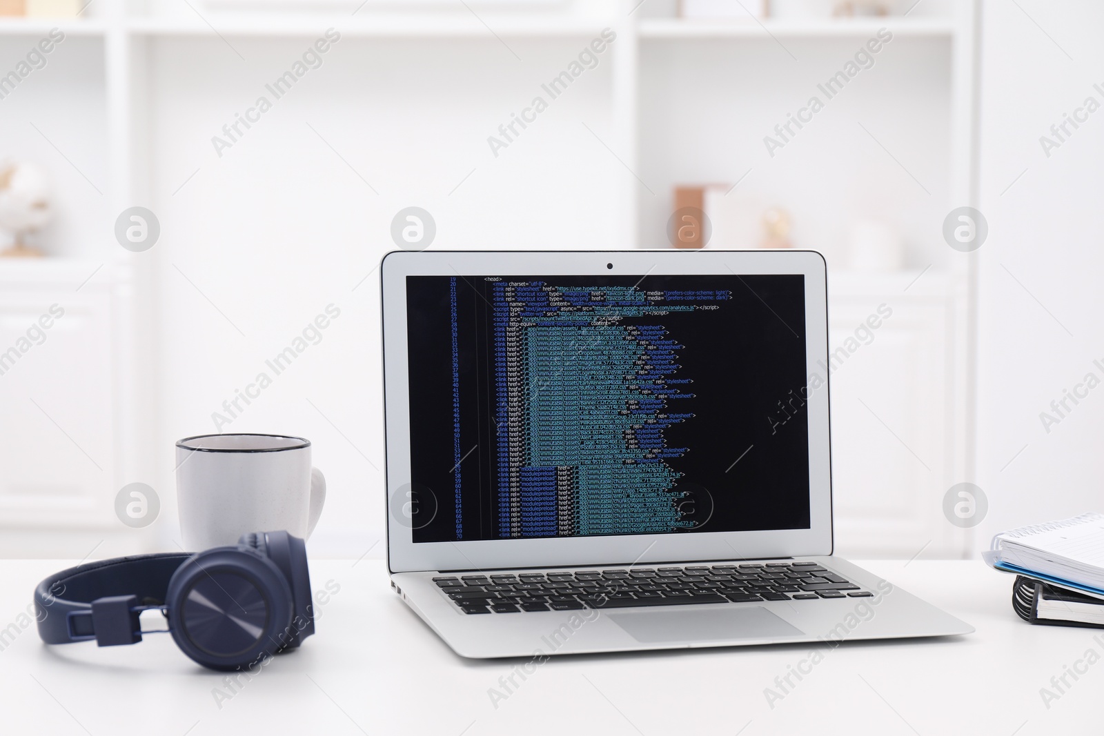 Photo of Programmer's workplace. Modern laptop with unfinished code, headphones and other supplies on white desk indoors