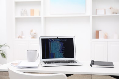 Photo of Programmer's workplace. Modern laptop with unfinished code, cup and notebooks on desk indoors