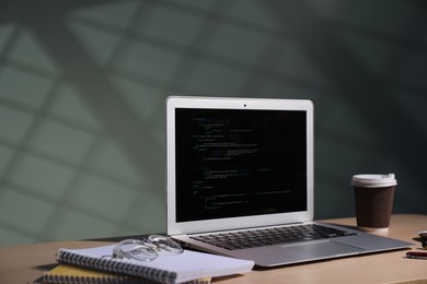 Photo of Programmer's workplace. Modern laptop with unfinished code, notebooks, glasses and paper cup of drink on wooden desk