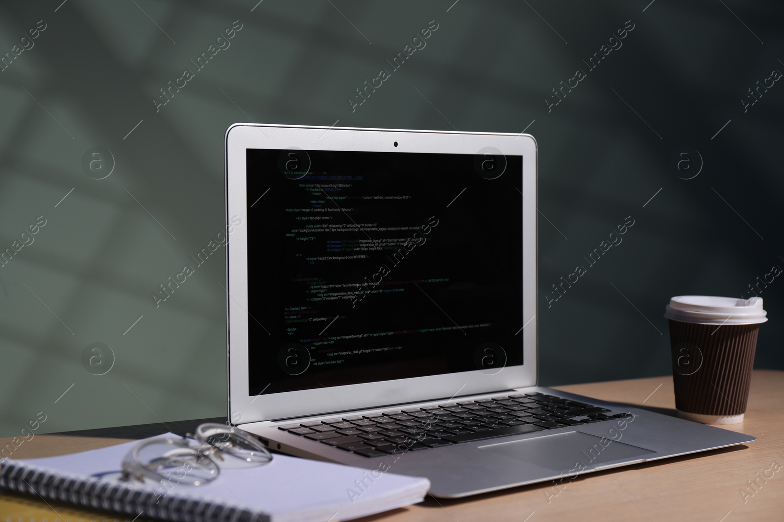 Photo of Programmer's workplace. Modern laptop with unfinished code, notebooks, glasses and paper cup of drink on wooden desk