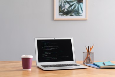 Photo of Programmer's workplace. Modern laptop with unfinished code, paper cup of drink and stationery on desk indoors