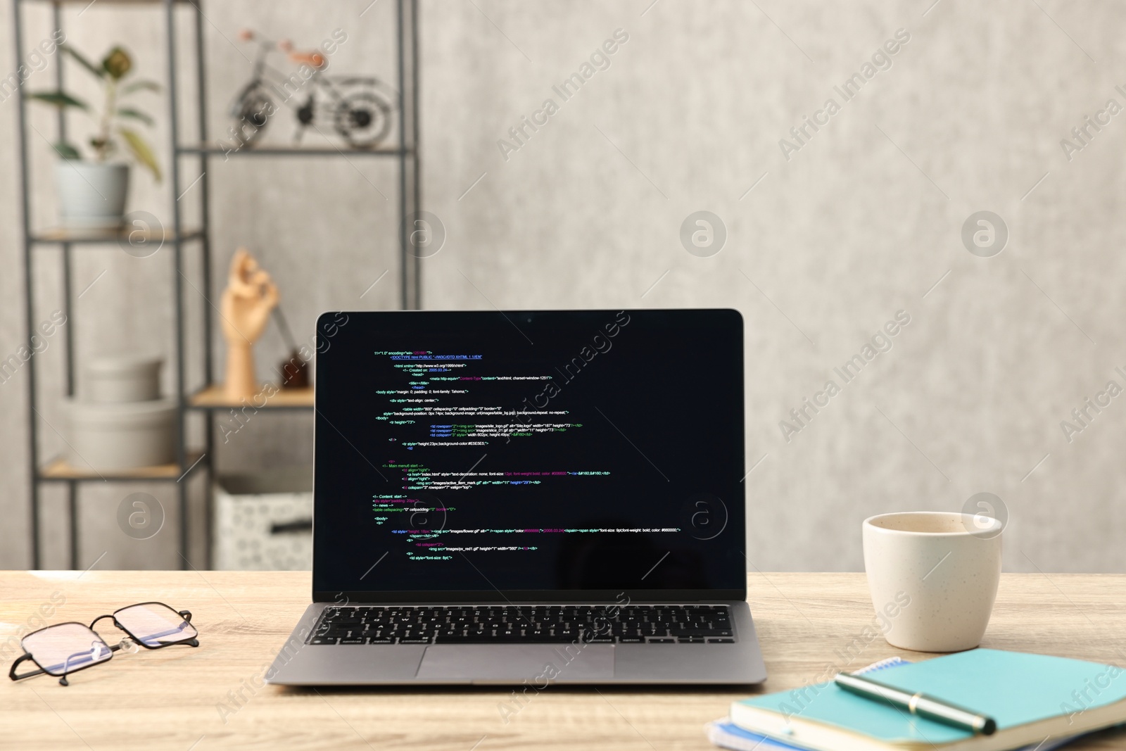 Photo of Programmer's workplace. Modern laptop with unfinished code, glasses cup and stationery on desk indoors