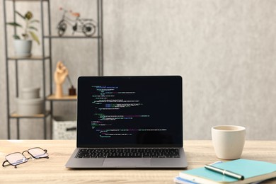 Photo of Programmer's workplace. Modern laptop with unfinished code, glasses cup and stationery on desk indoors