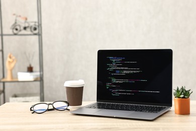 Photo of Programmer's workplace. Modern laptop with unfinished code, paper cup of drink, glasses and houseplant on desk indoors