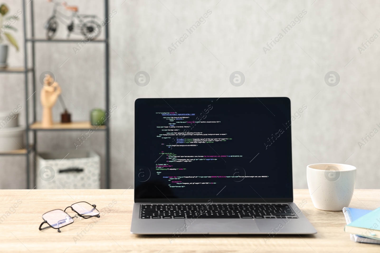 Photo of Programmer's workplace. Modern laptop with unfinished code, cup, glasses and stationery on desk indoors