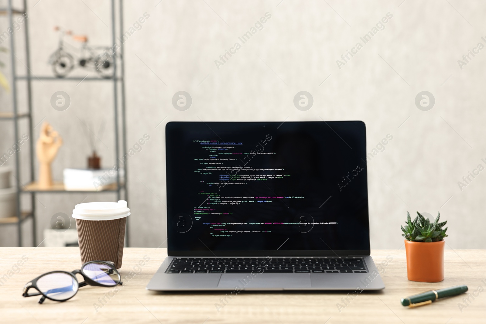 Photo of Programmer's workplace. Modern laptop with unfinished code, paper cup of drink, glasses and houseplant on desk indoors