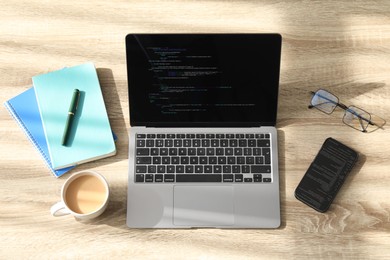 Photo of Programmer's workplace. Modern laptop with unfinished code, smartphone and other supplies on wooden desk, flat lay