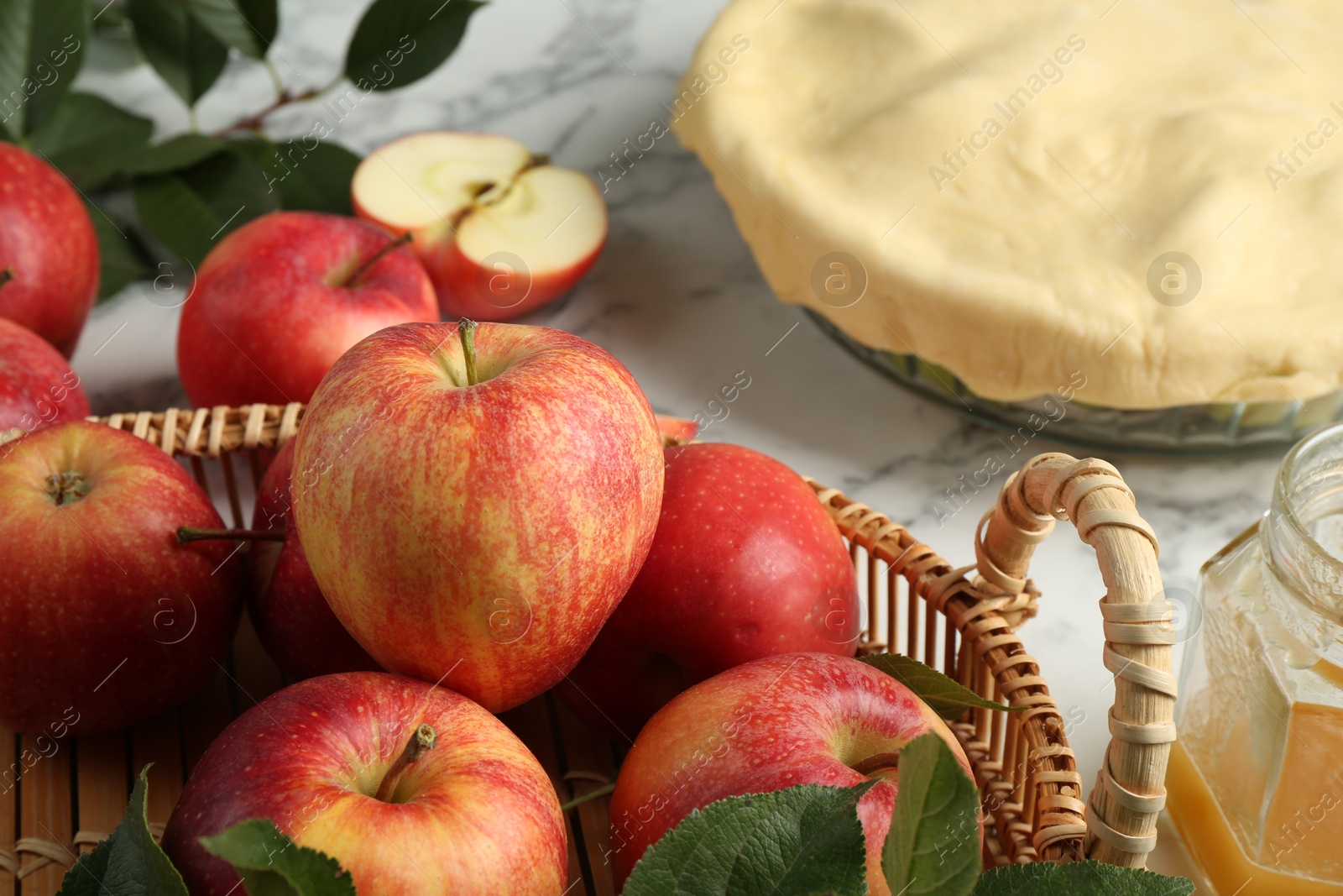 Photo of Wicker basket with fresh red apples and raw homemade pie on white marble table, closeup