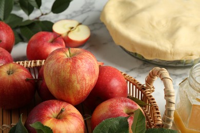 Photo of Wicker basket with fresh red apples and raw homemade pie on white marble table, closeup