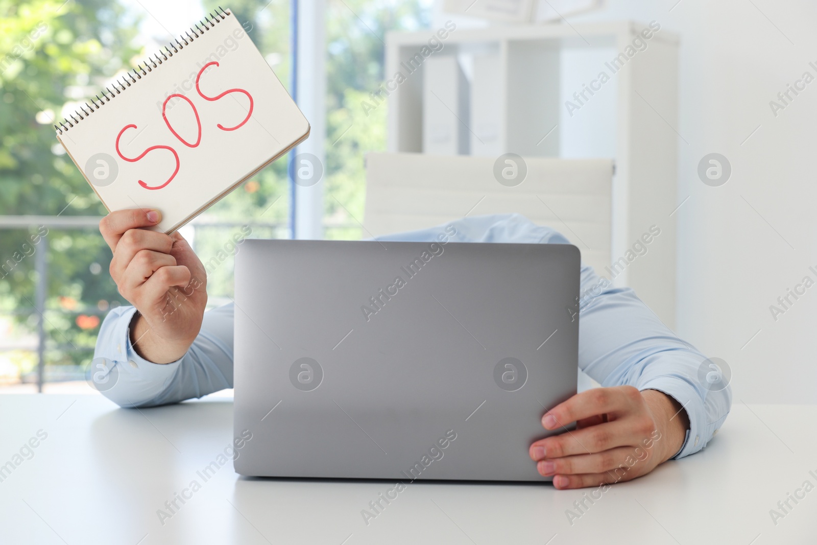 Photo of Man holding notebook with word SOS and hiding face behind laptop at table in office