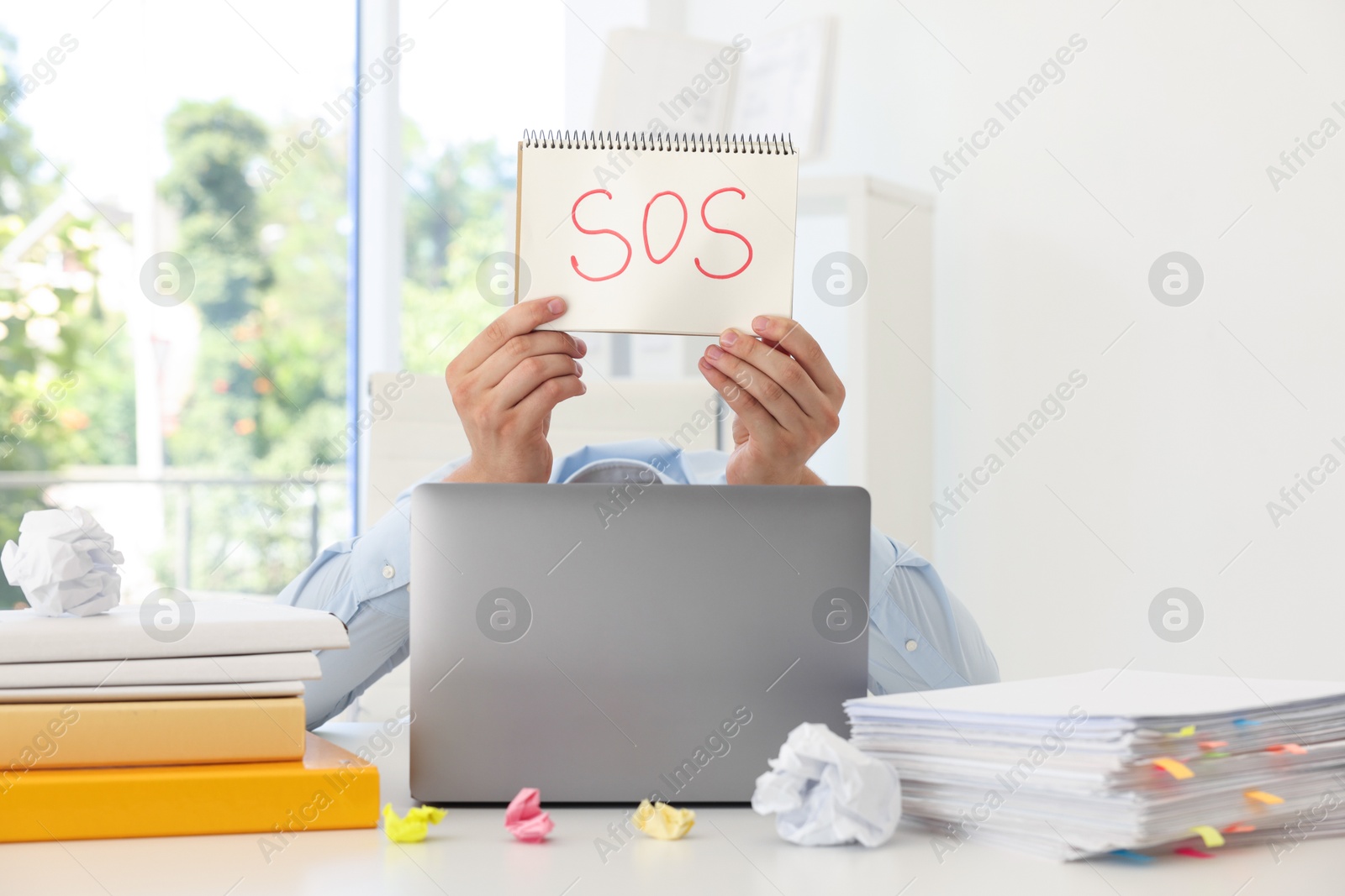 Photo of Man holding notebook with word SOS and hiding face behind laptop at table in office