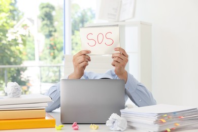 Photo of Man holding notebook with word SOS and hiding face behind laptop at table in office