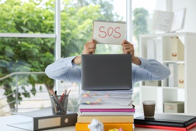 Photo of Man holding notebook with word SOS and hiding face behind laptop at table in office