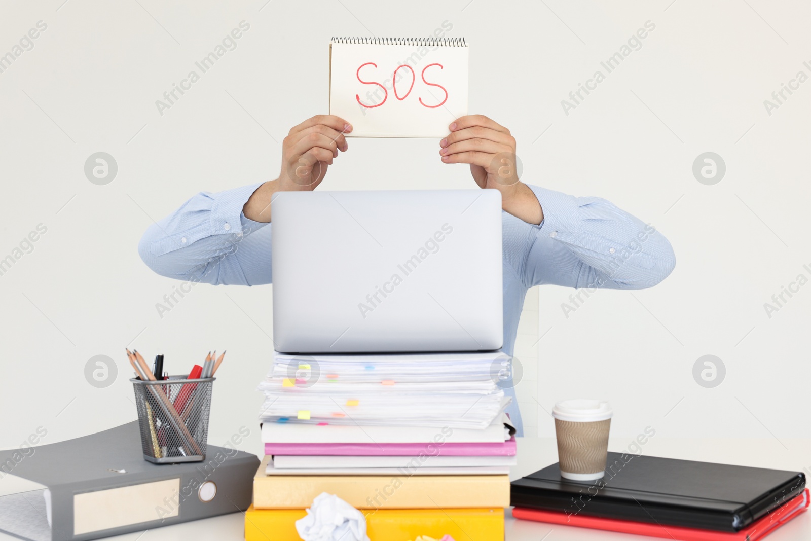 Photo of Man holding notebook with word SOS and hiding face behind laptop at table on light background