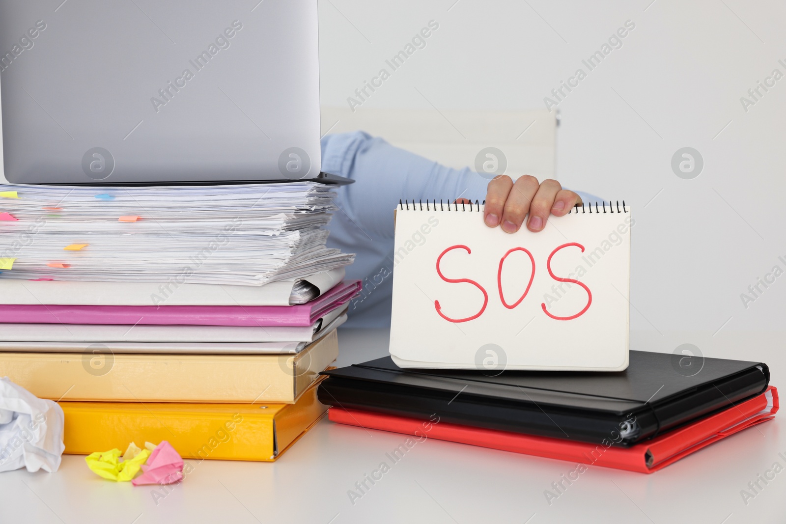 Photo of Man holding notebook with word SOS and hiding face behind laptop at table on light background