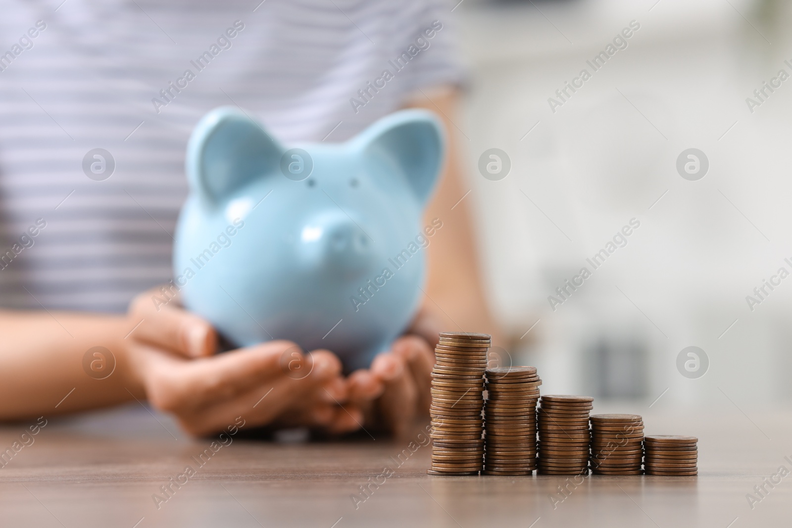 Photo of Woman with piggy bank at wooden table indoors, focus on stacks of coins