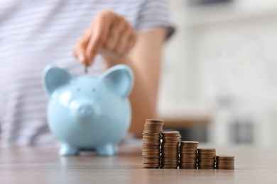 Woman putting coin into piggy bank at wooden table indoors, focus on stacks of coins